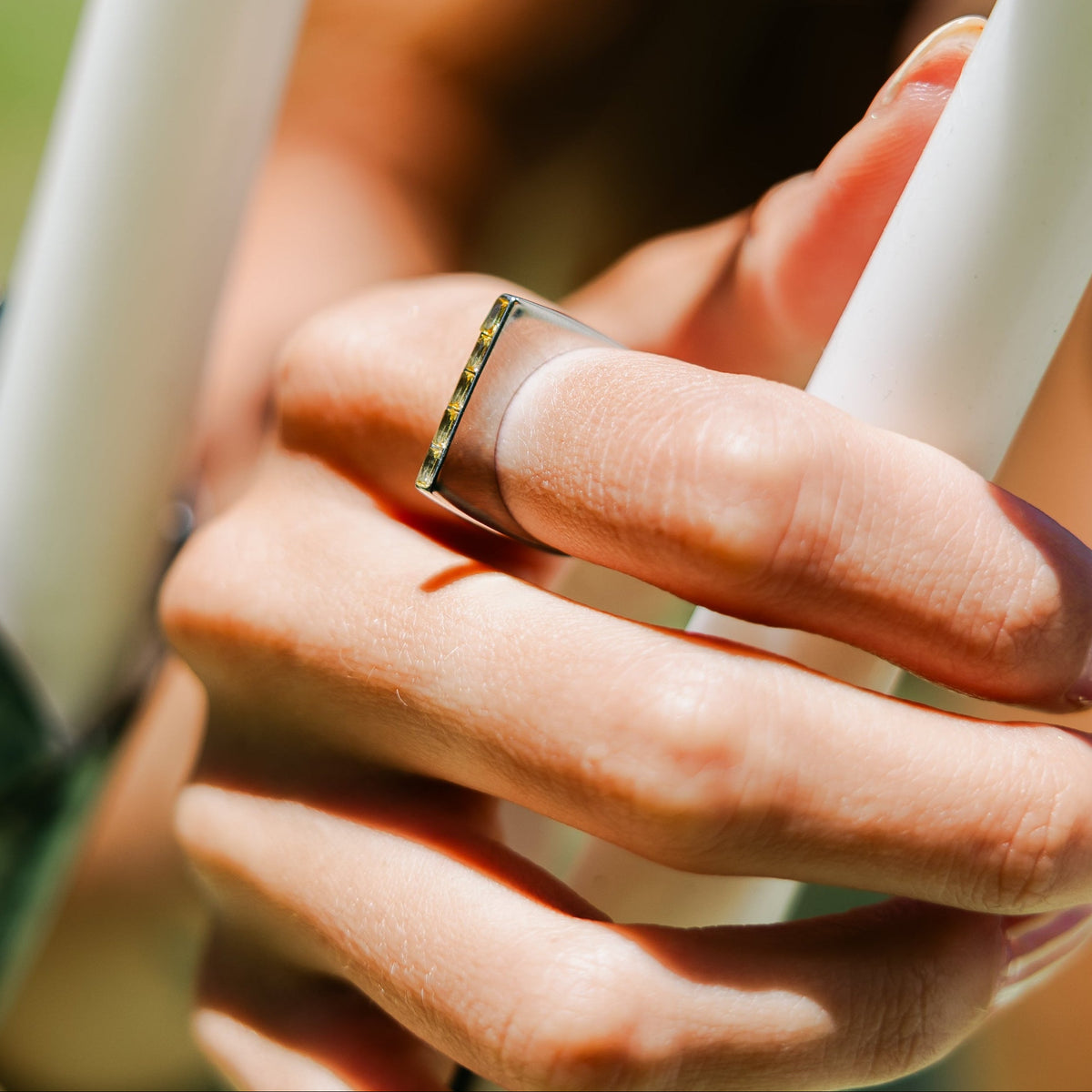 A close-up of a person's hand wearing a wide silver band ring with a textured, patterned gold-toned inlay on one side, holding a white pole outdoors.